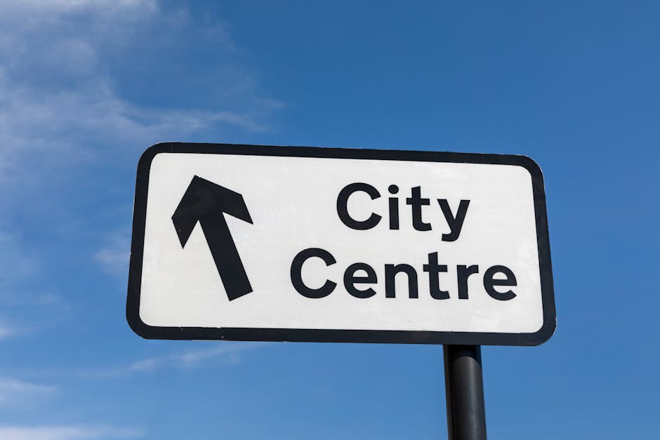 A rectangular road sign mounted on a black pole against a bright blue sky with some scattered clouds, indicating the direction towards the city centre with a large black arrow pointing straight ahead. The sign has a white background with black text that reads 'City Centre' in bold, clear font. It is positioned at an outdoor location, likely near a residential or commercial area, and is used to direct drivers or pedestrians along a planned route for urban navigation or house removals. The simplicity of the sign and the clear weather conditions suggest a typical daytime setting, useful for planning logistics in home relocation or furniture transport when moving through city areas, as seen by services like Man and Van Edgware.