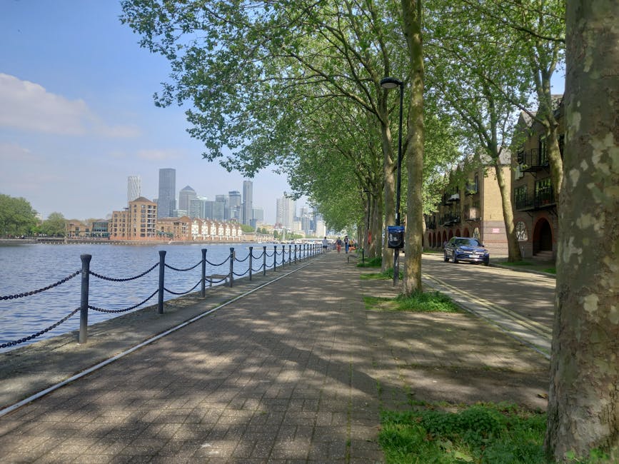 A paved riverside promenade lined with green-leaved trees casting shadows onto the pathway, which runs parallel to the water. On the right, there are modern residential buildings with balconies and cars parked along the curb. A street lamp, mailbox, and a few pedestrians are visible further along the path. The river on the left side features a black metal railing with decorative chains, separating the walkway from the water. In the distance across the river, a city skyline with tall office buildings and skyscrapers can be seen under a partly cloudy sky. This scene illustrates an urban house relocation or moving route with outdoor space, typical of a street used for furniture transport and packing and moving activities near residential and commercial areas, as would be seen in comprehensive house removals services by Man and Van Edgware.