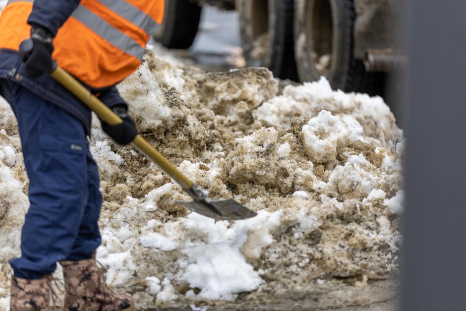 A person wearing a high-visibility orange and black jacket, blue work trousers, and rubber boots is using a shovel to clear a large pile of dirty, mixed snow and slush from the ground outside a property. The snow appears wet and partially melted, with brown dirt and debris mixed in. In the background, a vehicle with large tires is parked on the pavement, which is adjacent to a doorway leading into a building. The scene takes place during daylight hours with natural light illuminating the area. This image captures the process of clearing snow, which may be a preparatory step before the loading and carrying of household items or furniture during a home relocation or moving service performed by [COMPANY_NAME], such as those offered on the Edgware High Street removals guide for tight access jobs, EDGWARE.