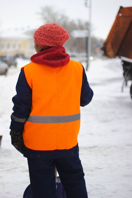 A person wearing a bright orange high-visibility safety vest over a dark coat, with a red knitted hat, standing outside on a snowy pavement. The individual is facing away from the camera and appears to be involved in a house removal or moving process, possibly coordinating or overseeing the loading of items. In the background, there is a large brown truck with its rear liftgate raised, indicating it is used for transporting furniture and boxes during a home relocation. The scene suggests cold weather conditions, with snow covering the ground and a cloudy sky overhead. This image exemplifies the logistics and careful handling involved in furniture transport and packing for house removals, which Man and Van Edgware provides as part of their professional removal services.