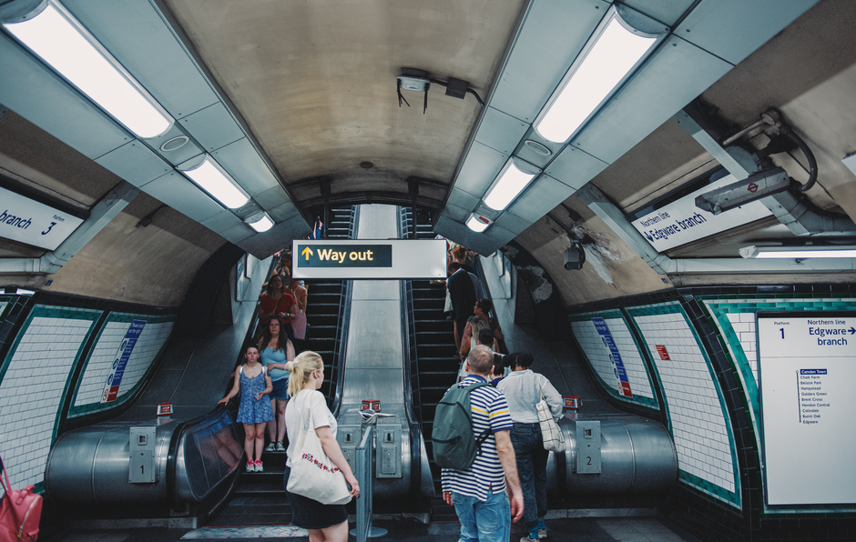 A busy underground train platform filled with passengers walking towards and away from arriving trains, with some individuals carrying backpacks and personal belongings. The platform features a digita
